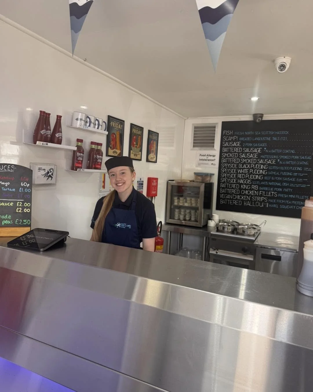 A smiling female staff member in a uniform standing behind a counter in a restaurant or cafe, with a menu board on the wall and kitchen equipment in the background.
