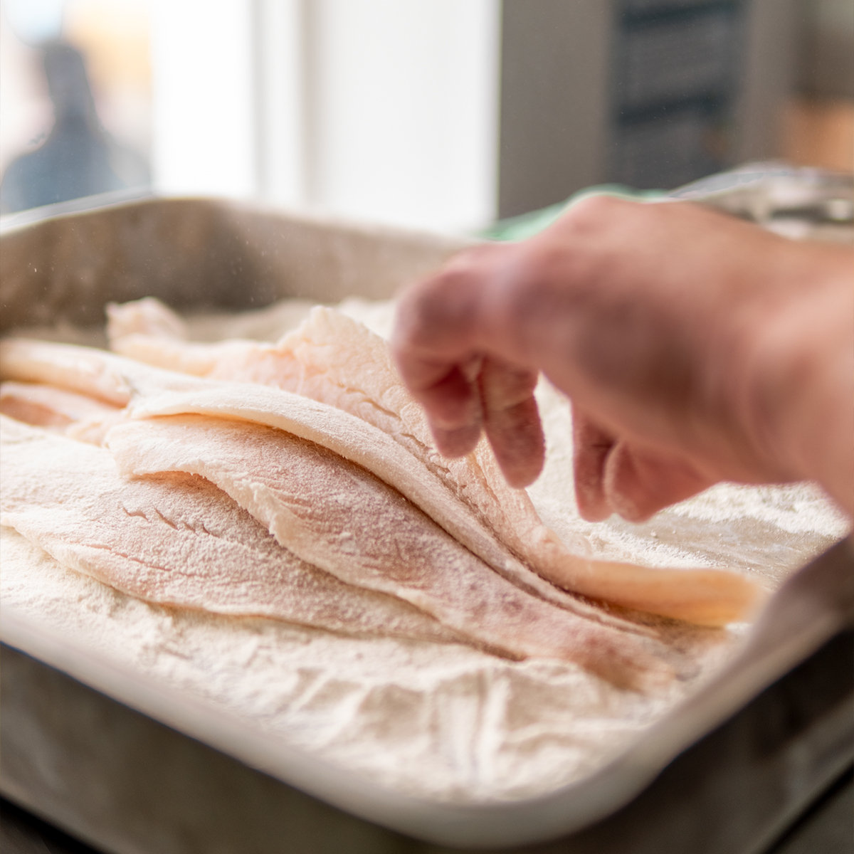 Person handling uncooked fish fillets on a floured surface.