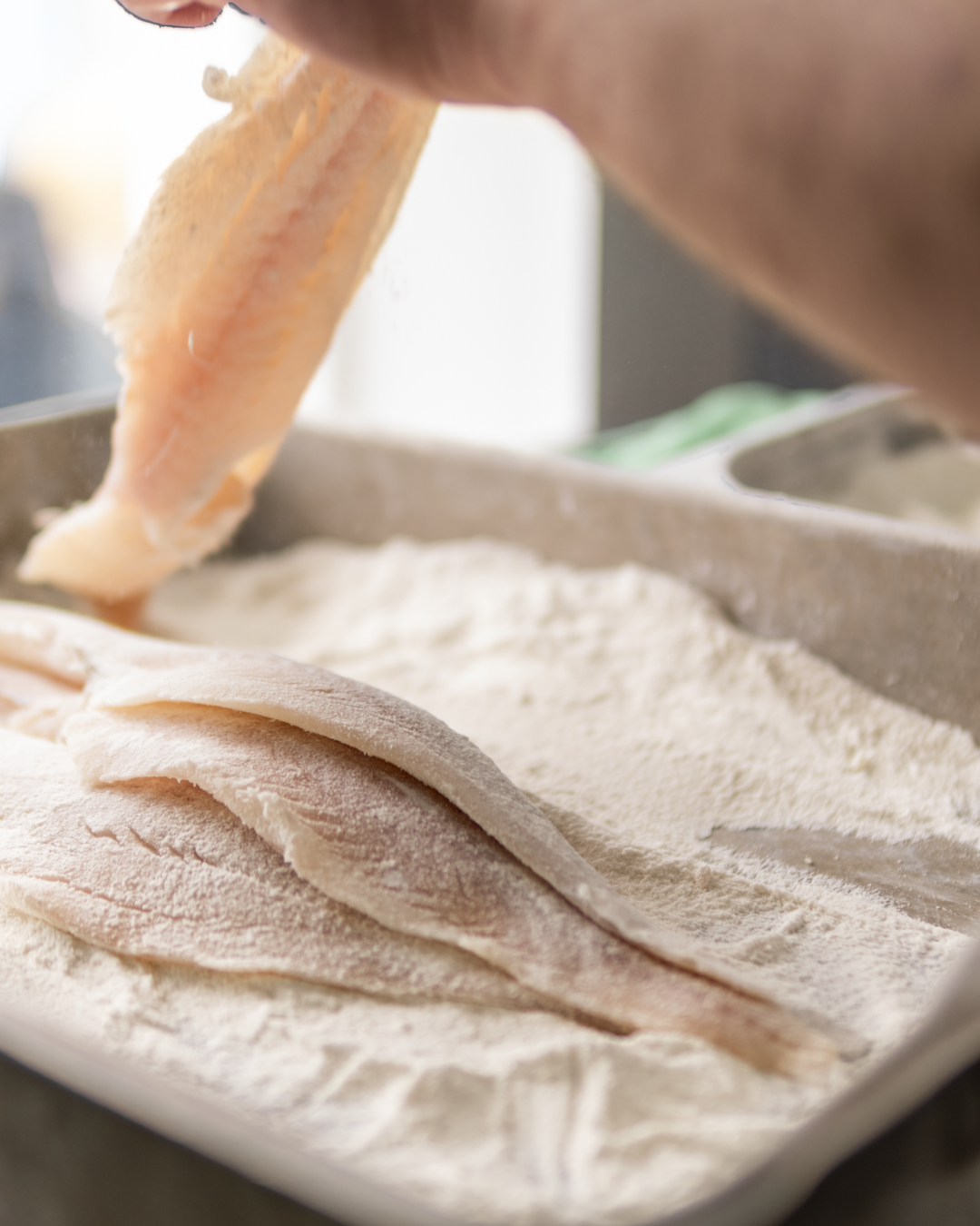 Person coating raw chicken fillets with flour on a baking sheet.