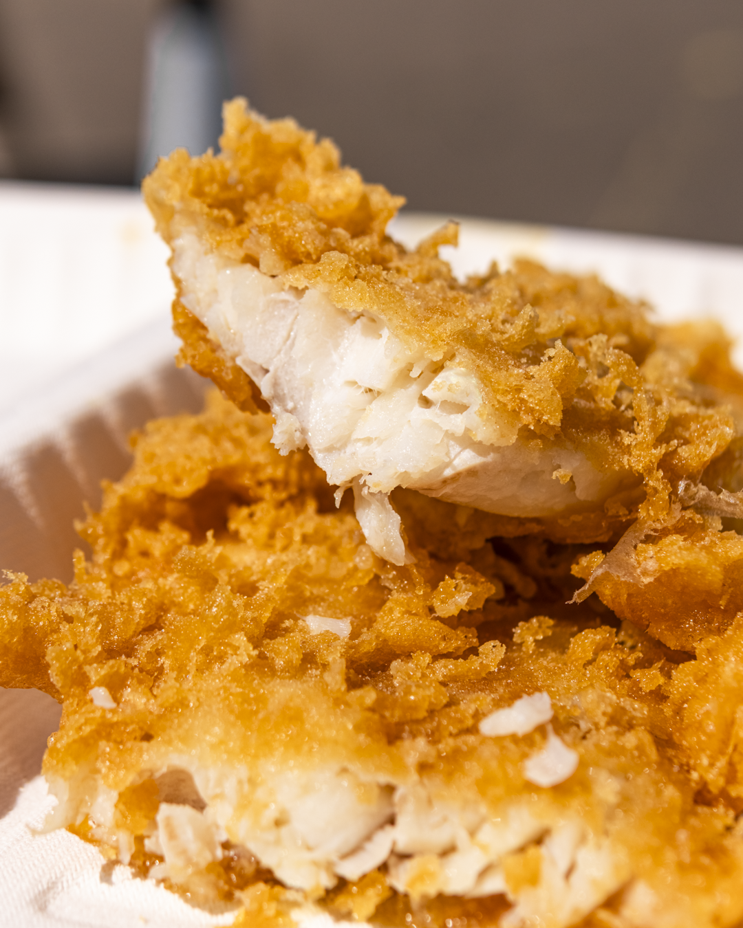 Close-up of crispy, golden fried fish fillet with a flaky white interior, placed on a white paper tray.