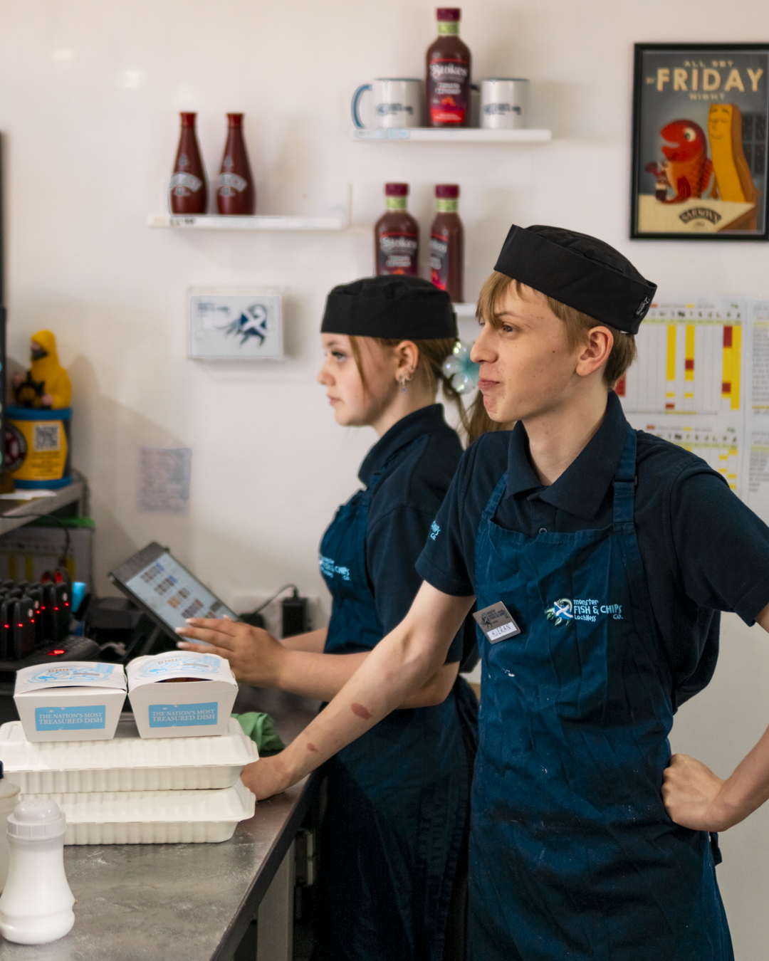 Two workers in blue shirts and black hats working at a seafood restaurant. One is using a touch screen, and the other is standing with her hands on her hips. There are ketchup bottles and boxes of seafood on the counter.