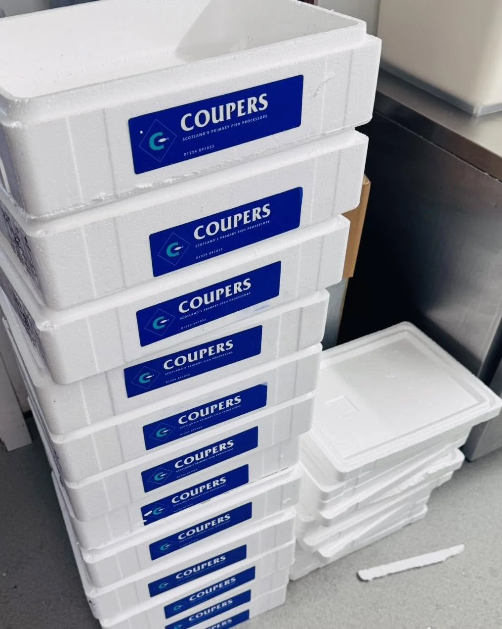 Stacked white insulated containers with blue labels reading "Couper's," used for Scottish primary fish processing, on a gray floor with additional containers nearby.