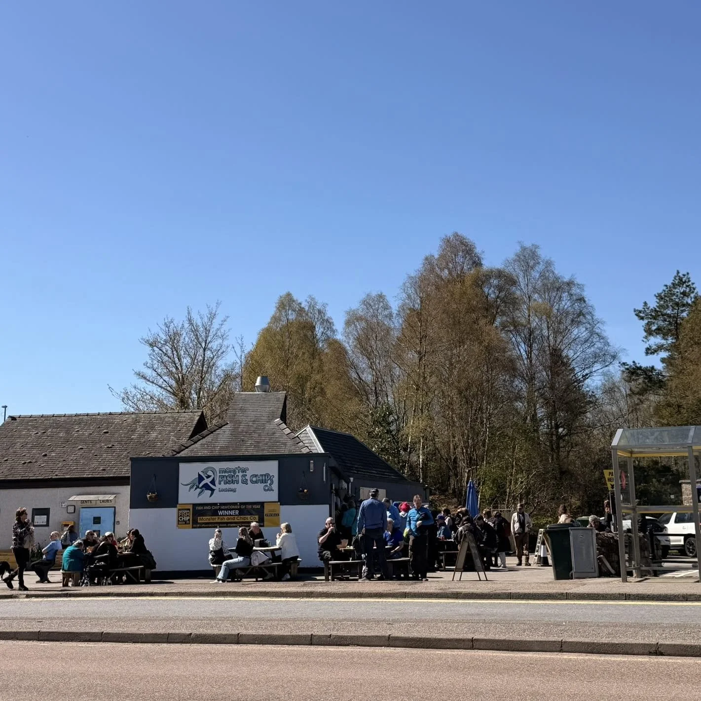 People sitting outside at tables in front of a building with a sign that reads 'Monster Fish & Chips Co.' under a clear blue sky.