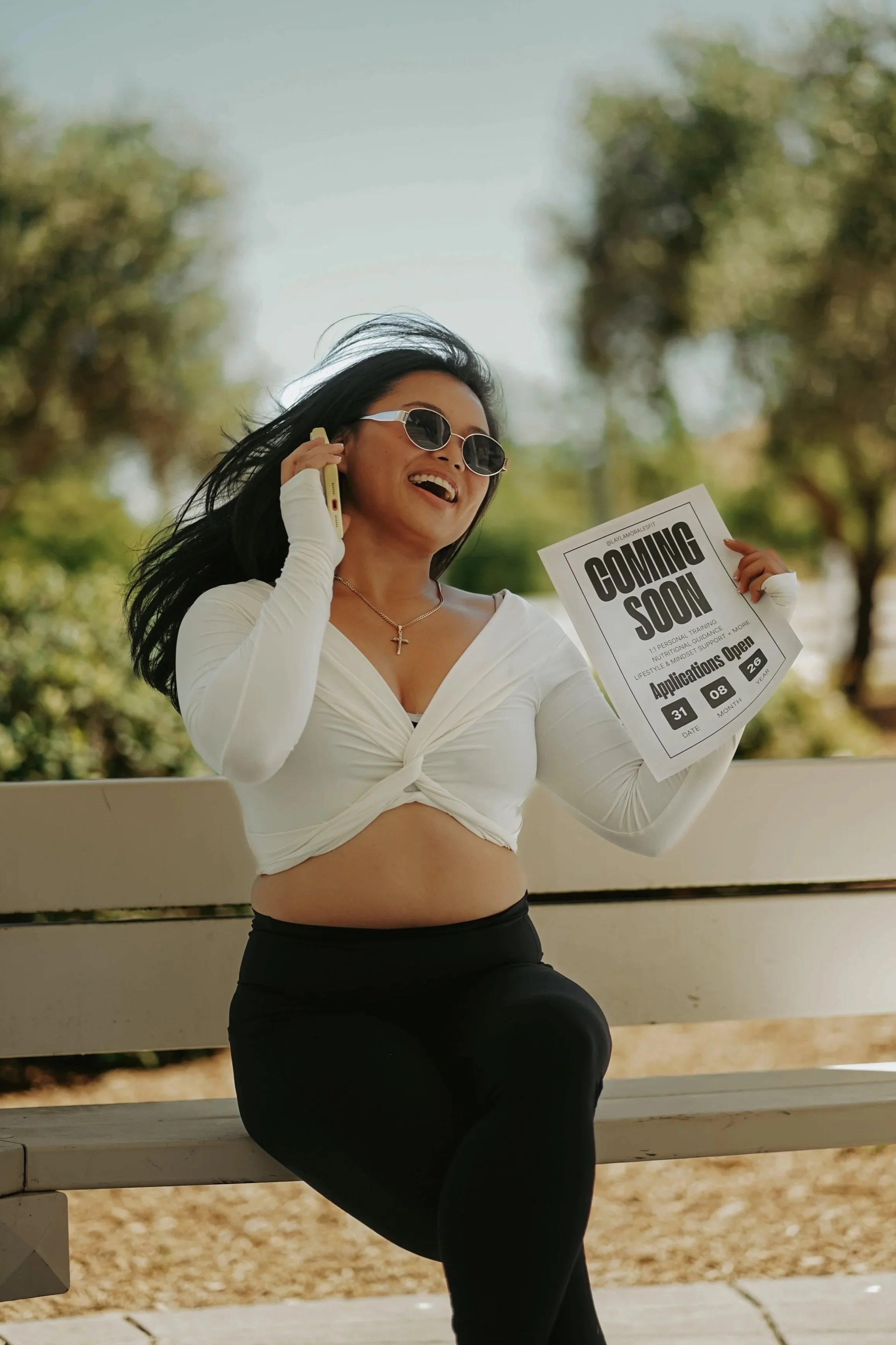 Woman with dark hair wearing sunglasses and a white long-sleeve top, holding a phone to her ear and a "Coming Soon" sign, seated on a bench outdoors, with blurred trees in the background.