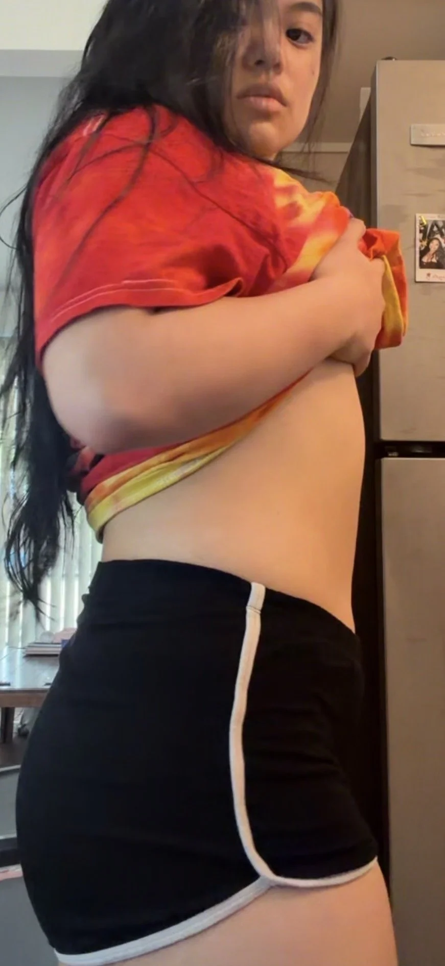A young woman lifting her shirt to reveal her midriff, standing in a room with a refrigerator in the background.