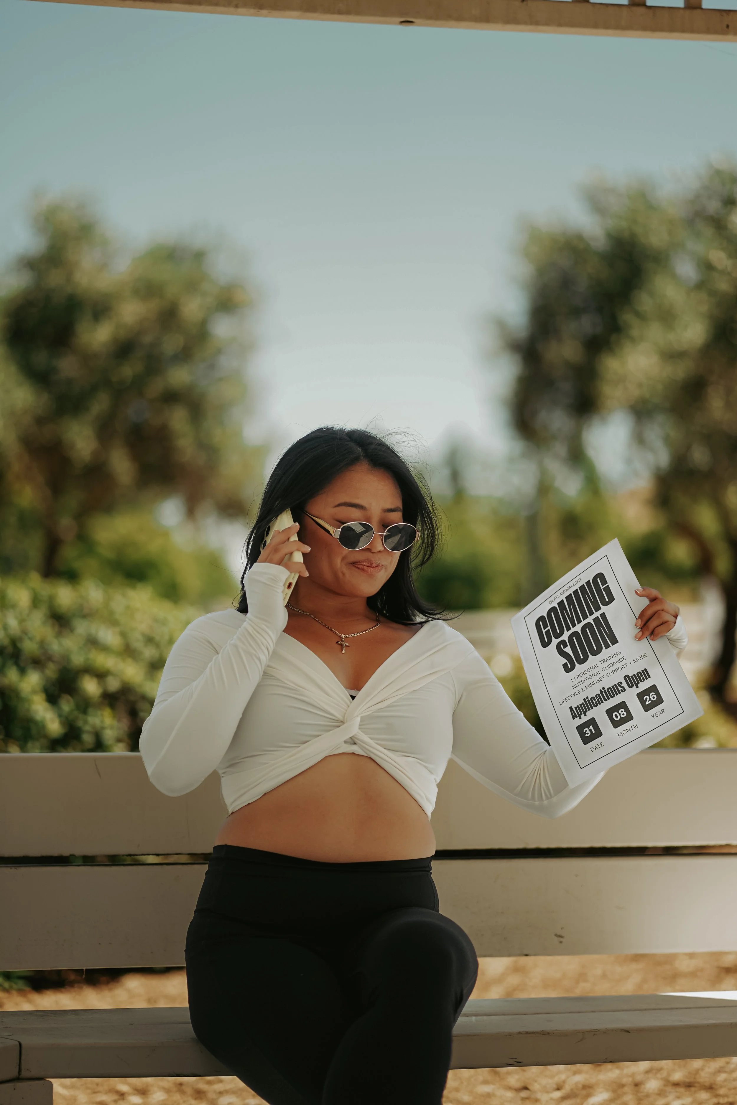 A woman sitting on a bench outdoors, talking on a cellphone, holding a printed application form, wearing sunglasses, a white knotted long-sleeve top, and black pants, with trees and clear sky in the background.