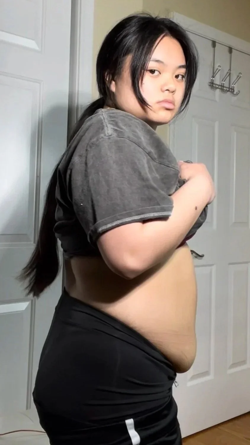 A young woman standing in a room with a closed white closet door, holding her shirt to reveal her stomach, looking at the camera with a neutral expression.