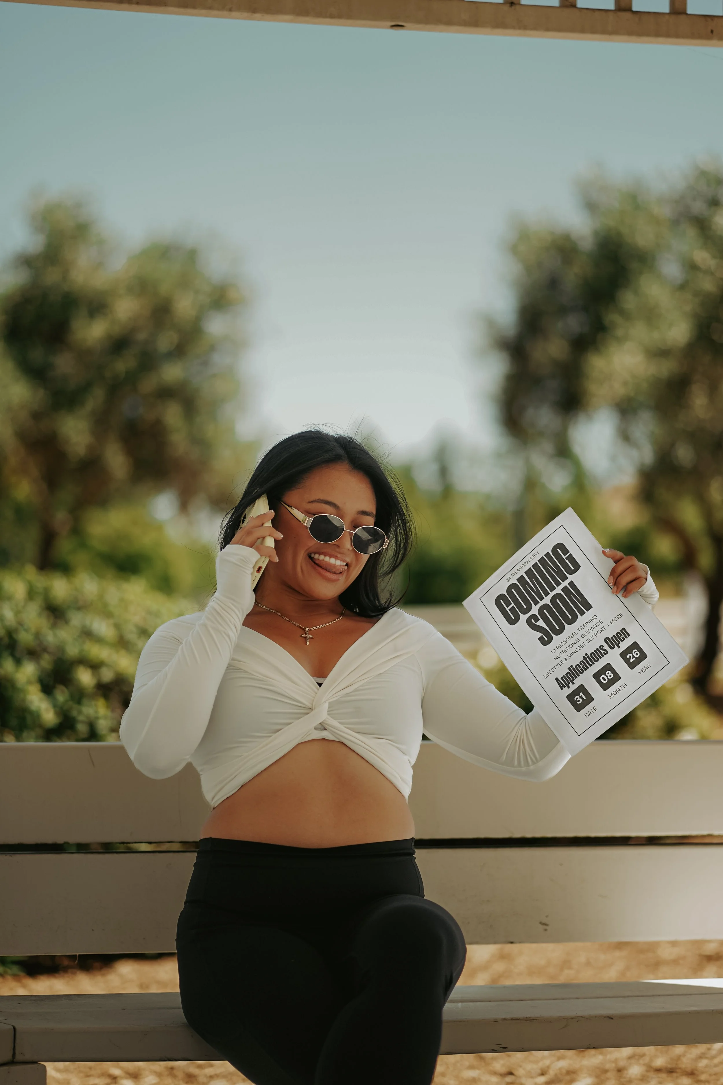 Woman with long dark hair, wearing sunglasses, white long-sleeve top, and black pants, holding a paper with 'Coming Soon' text and talking on a phone outdoors with trees in the background.