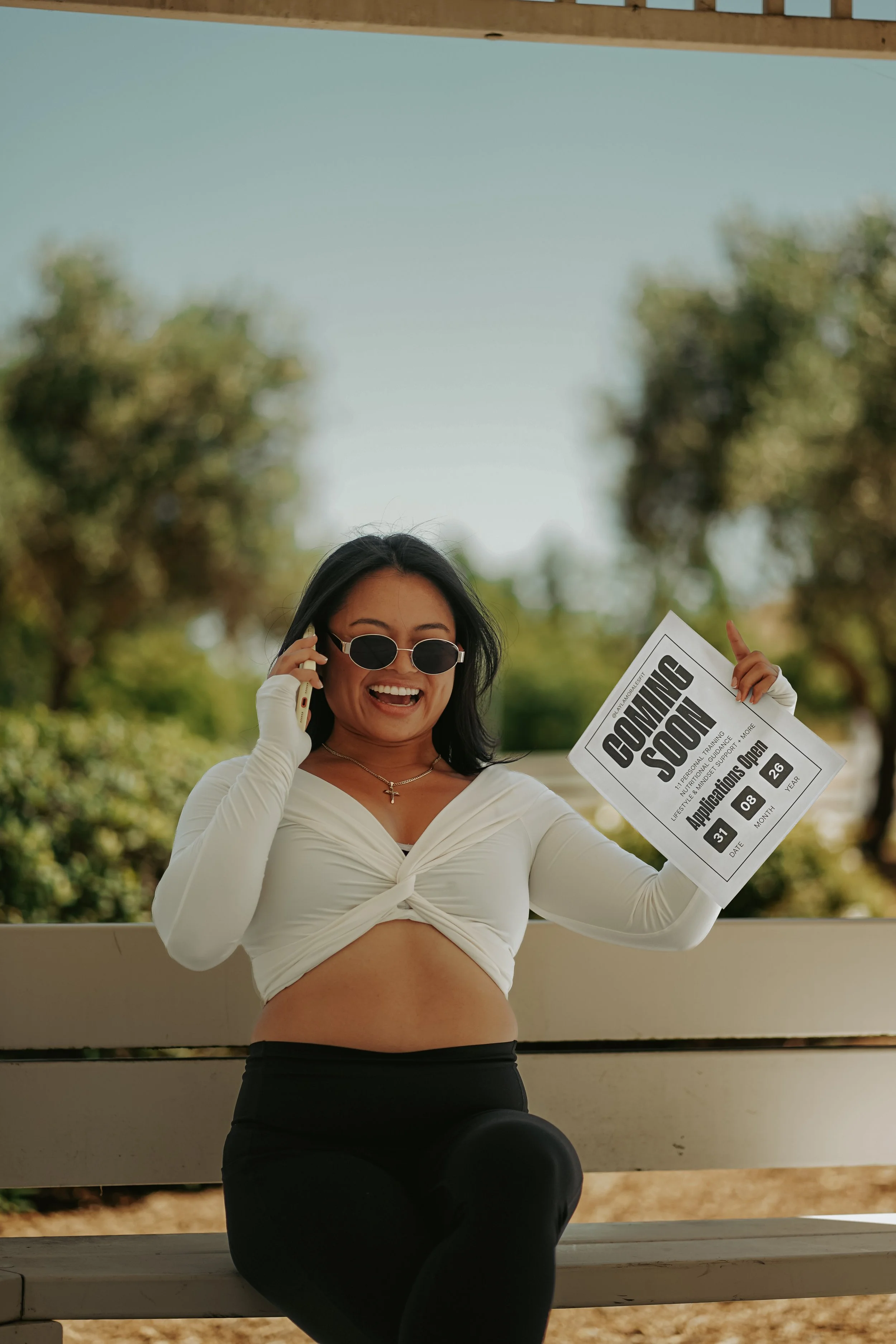 A woman smiling and talking on her cellphone, holding a document that says 'Coming Soon' and showing her tongue with sunglasses, outdoors with trees in the background.