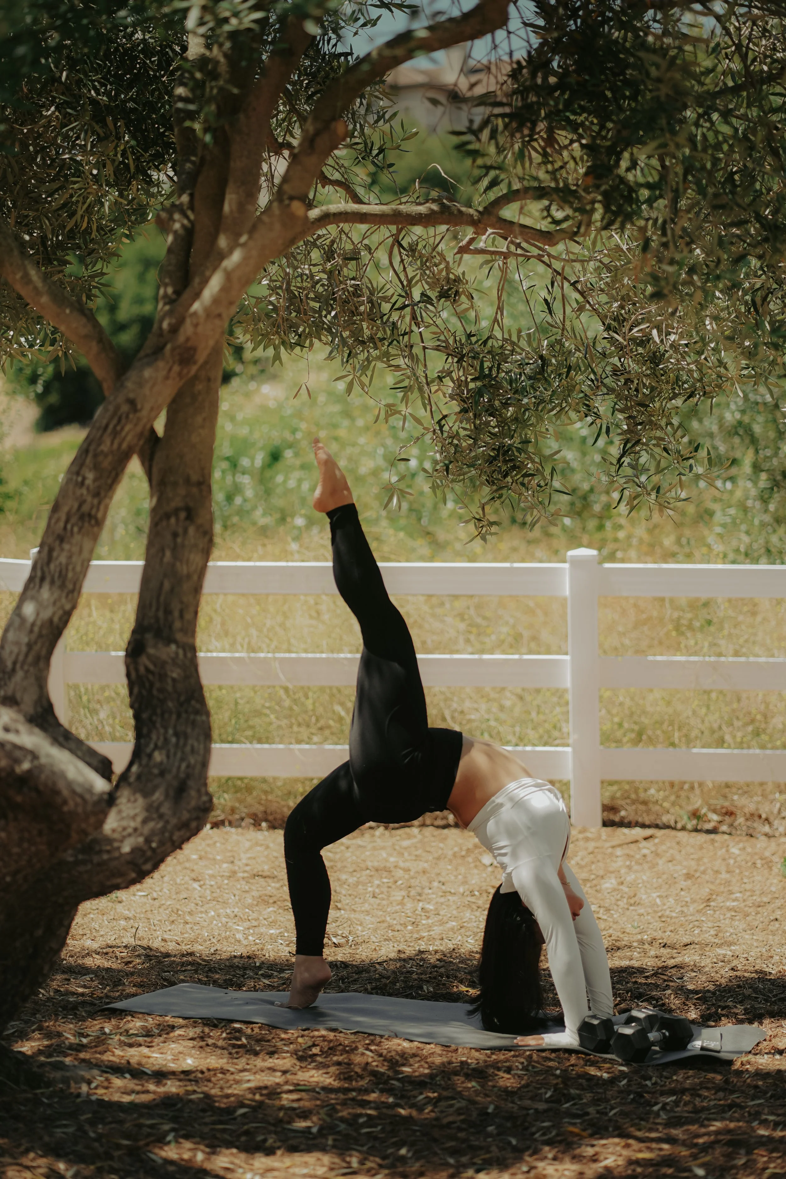 A woman practicing yoga outdoors in a handstand pose with her legs straight up against a tree, on a yoga mat, wearing workout clothes and a white head covering, in a sunny garden with trees and a white fence.