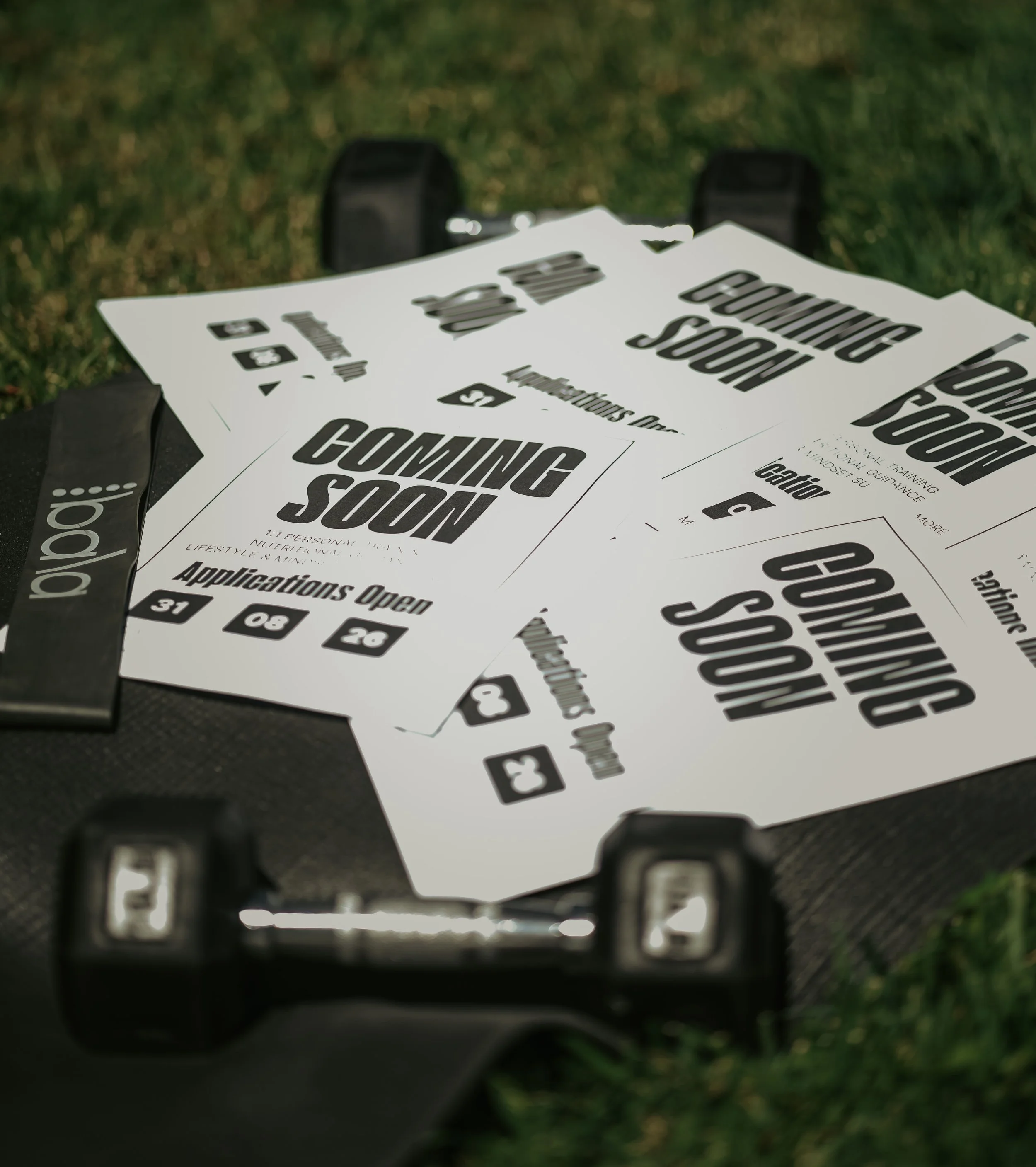 Close-up of a poker or application open ticket with bold black text that says "COMING SOON" and additional informational text. The paper rests on green grass with a black object and part of a metallic structure in the foreground.