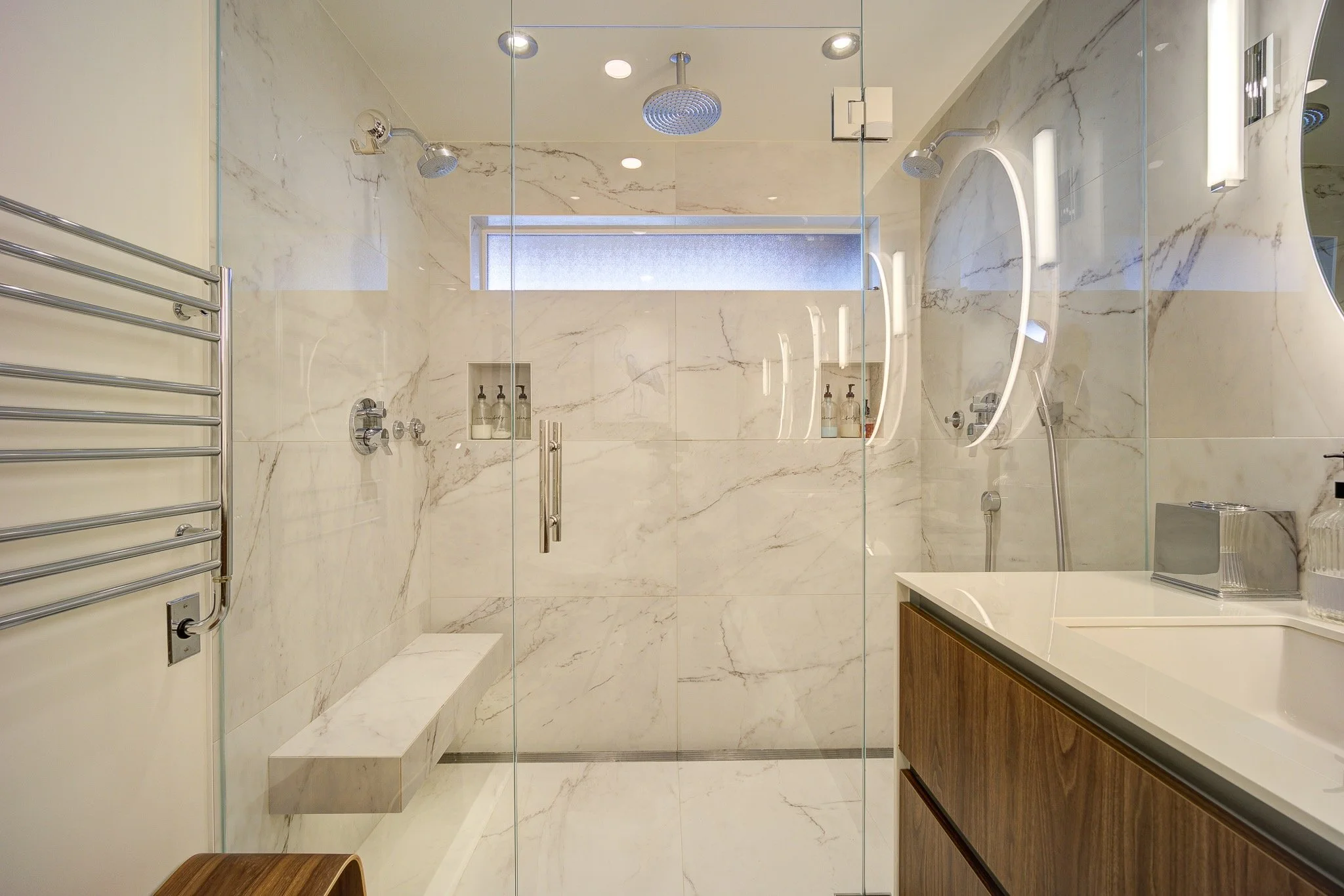 Modern walk-in shower with marble walls and a built-in marble bench. Dual shower heads and a sliding glass door. Shower products in a niche, minimalist and sleek design.