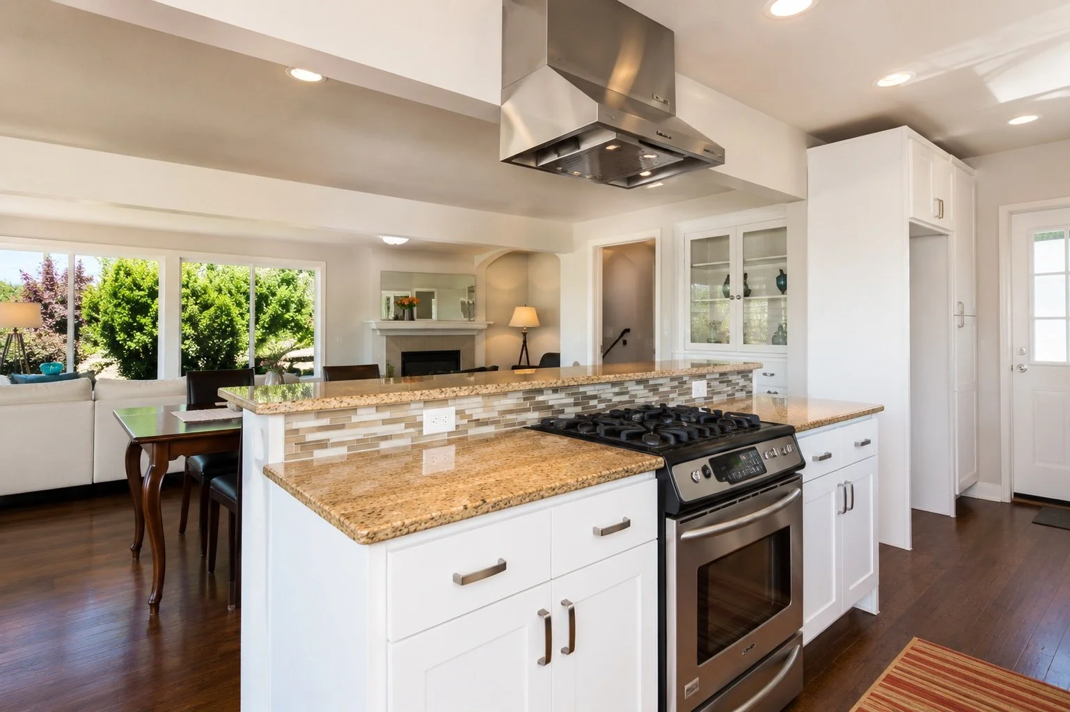Modern kitchen with granite countertops, white cabinets, stainless steel oven and stove, glass tile backsplash, and open view to living and dining areas with large windows and hardwood floors.
