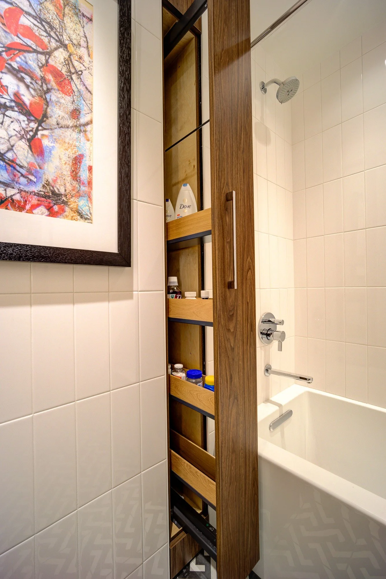 The image shows a bathroom with white tiled walls, a bathtub with a showerhead, a wooden cabinet with open shelves containing various toiletries, and a framed colorful abstract painting on the wall.