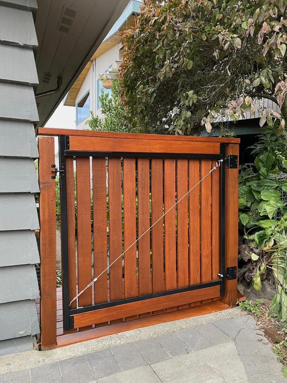 A wooden and metal gate with a diagonal support wire installed between posts. The gate is attached to a neighboring house or shed with gray siding, and there are green plants and bushes around it.