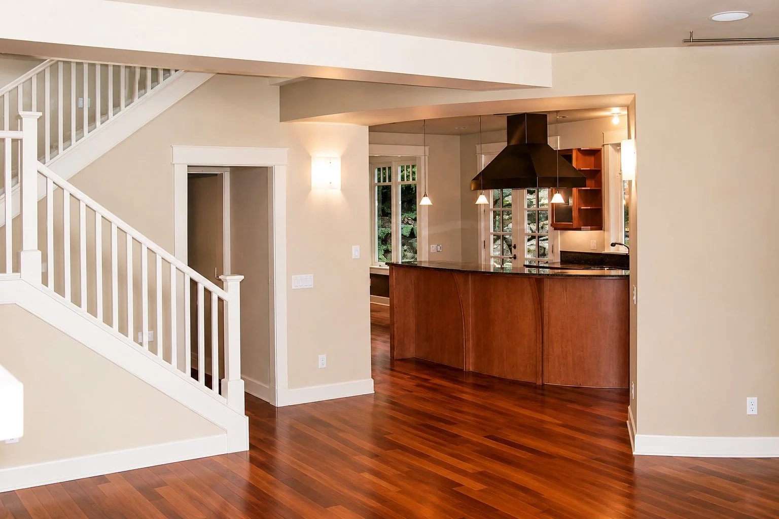 Open-concept living space with a staircase, hardwood floors, and a kitchen area with a black range hood and pendant lights.