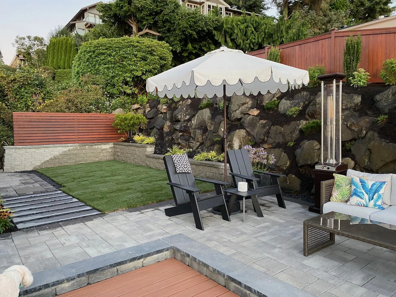 Backyard patio with two black Adirondack chairs and an umbrella, a white outdoor sofa with colorful pillows, patio heater, paved flooring, small lawn, stone retaining wall, greenery, and a wooden fence.