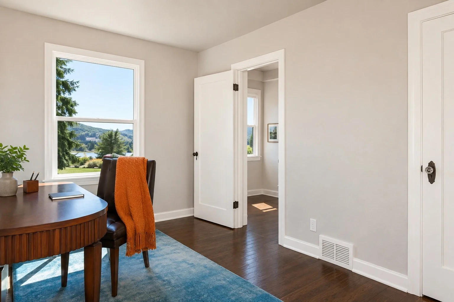 Interior view of a room with a window showing a scenic outdoor landscape, a wooden table with a chair and an orange throw blanket, and a partly open door leading to another space.