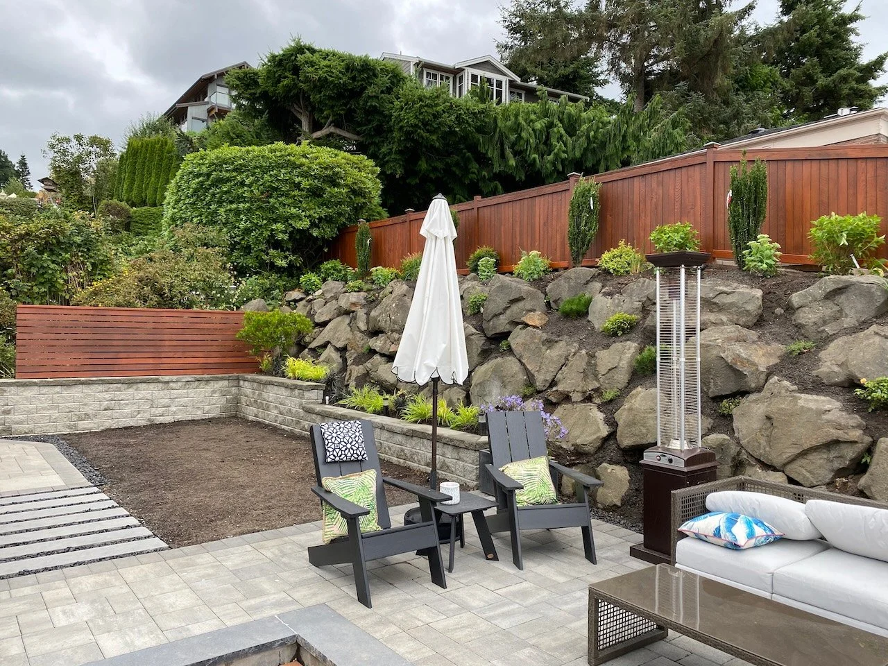 Backyard patio with two chairs, a table, a white sofa, a patio heater, a closed umbrella, and a landscaped rock wall with plants and trees.