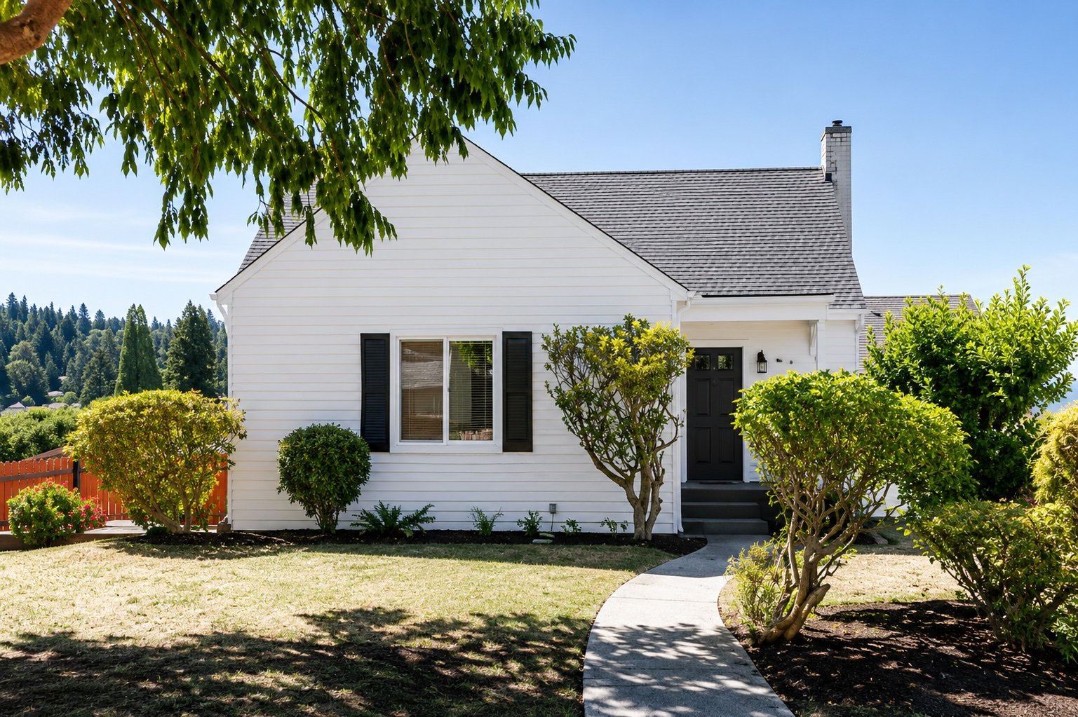 Front view of a white house with black shutters, surrounded by green bushes and trees, under a clear blue sky.