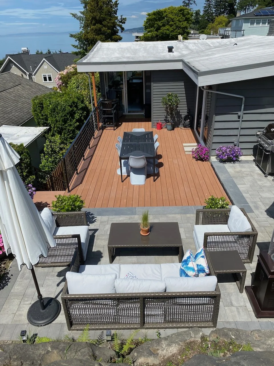 Top-down view of a backyard patio with outdoor furniture, including a sofa, chairs, a coffee table, and a dining table with chairs, surrounded by greenery and overlooking a body of water.