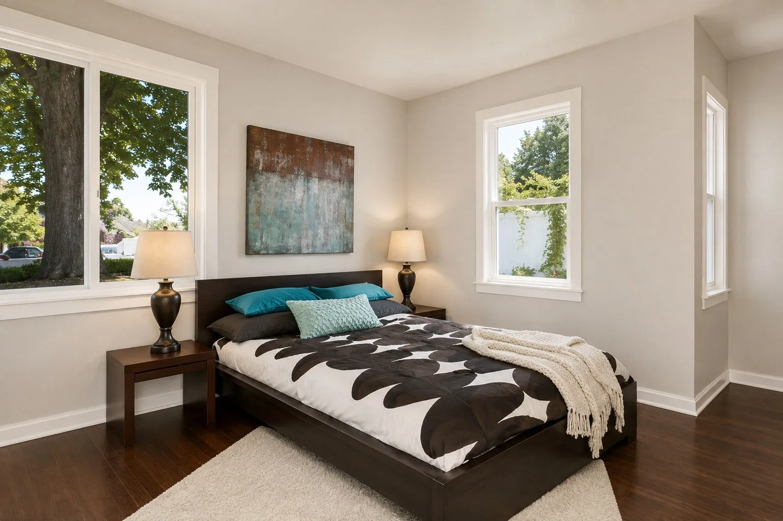 A modern bedroom with a dark wood bed, black and white floral bedding, teal and black pillows, and a cream knit throw. Flanked by two matching nightstands with lamps. Four windows show green trees outside.