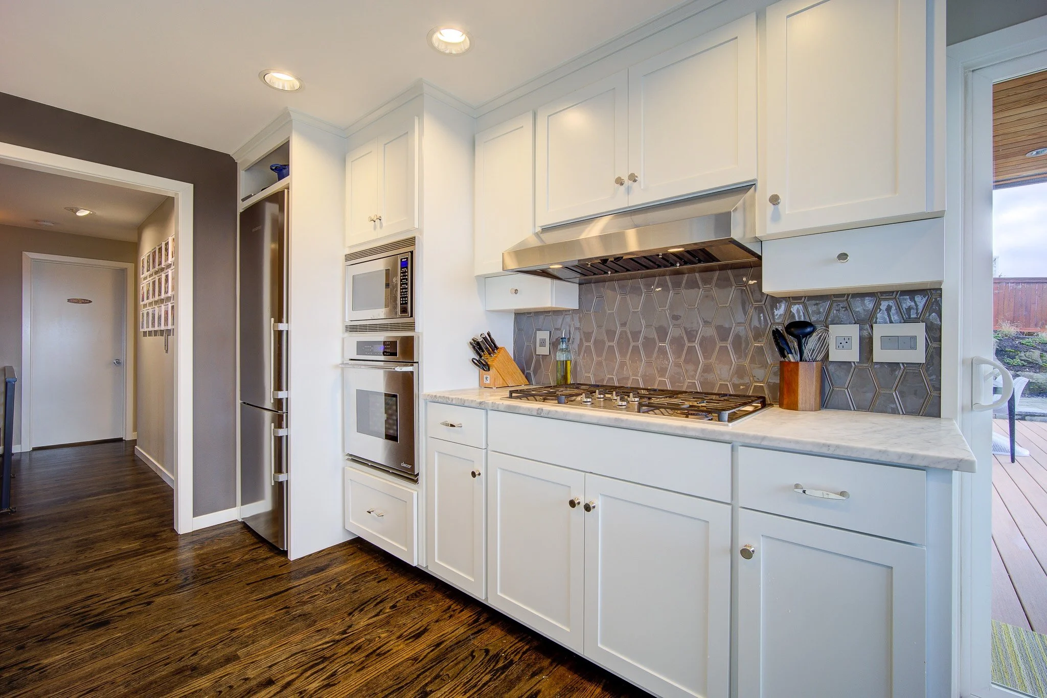 A modern kitchen with white cabinets, a marble countertop, stainless steel appliances, brown geometric backsplash, and a view of an outdoor deck.