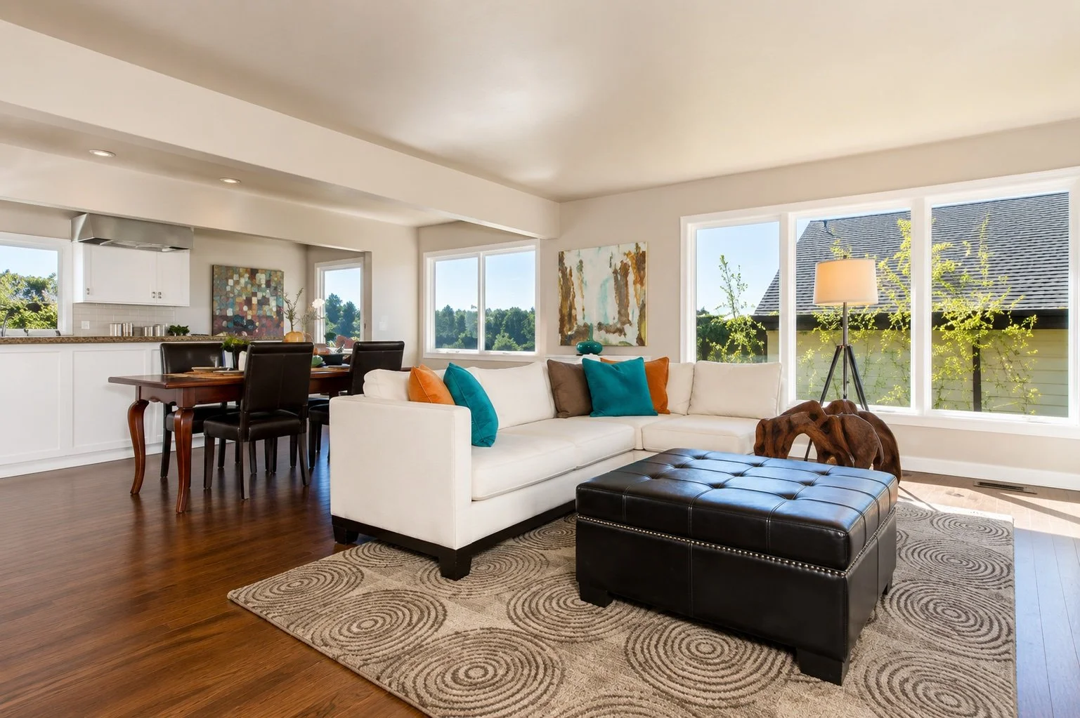 Bright living room with white sectional sofa, colorful throw pillows, a large window, artwork on the wall, wooden coffee table, and a black leather ottoman.