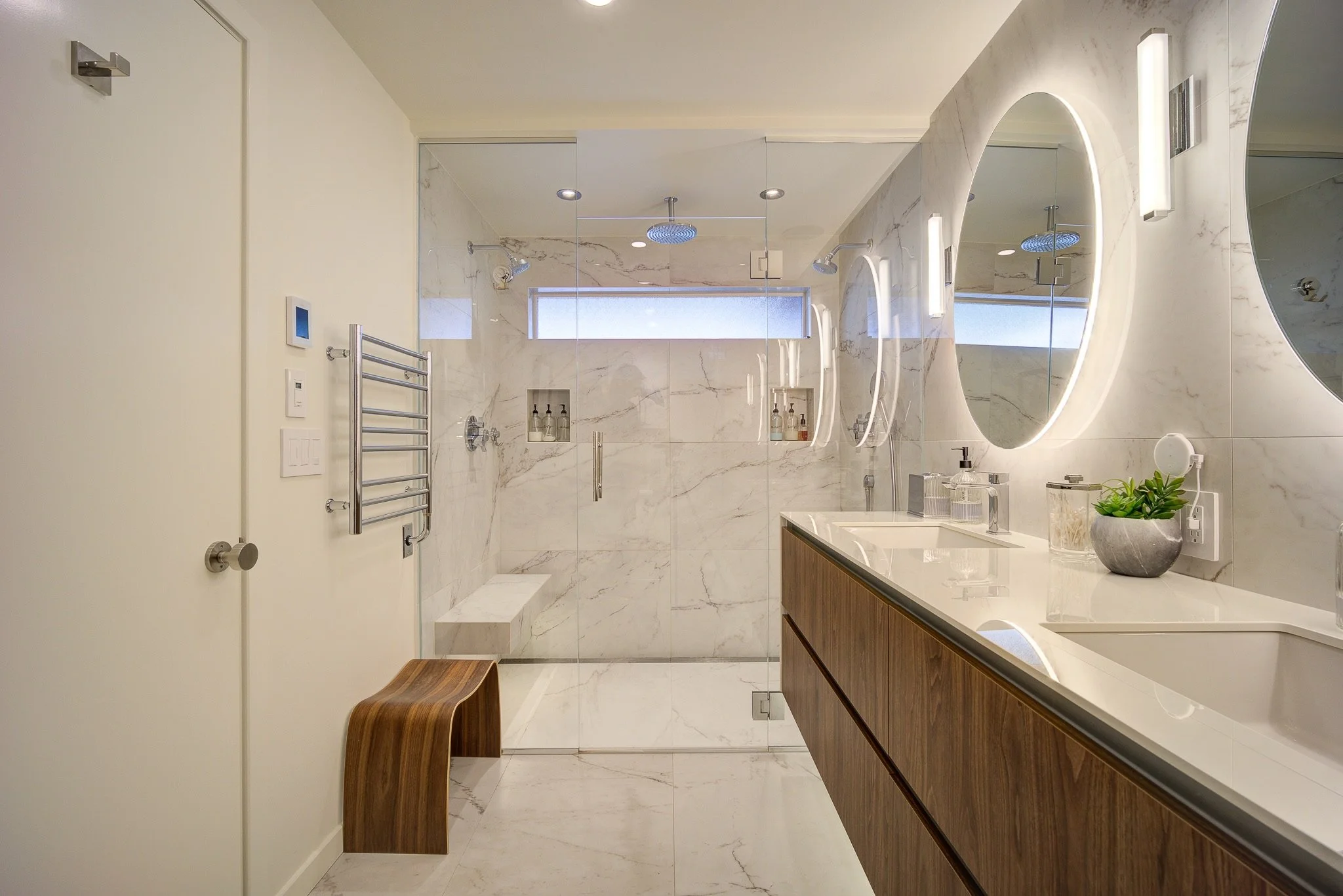 Modern bathroom with marble walls, ships vanity, and illuminated mirror, featuring a shower area with glass door, bench, and built-in shelves, along with decorative plants on the counter.