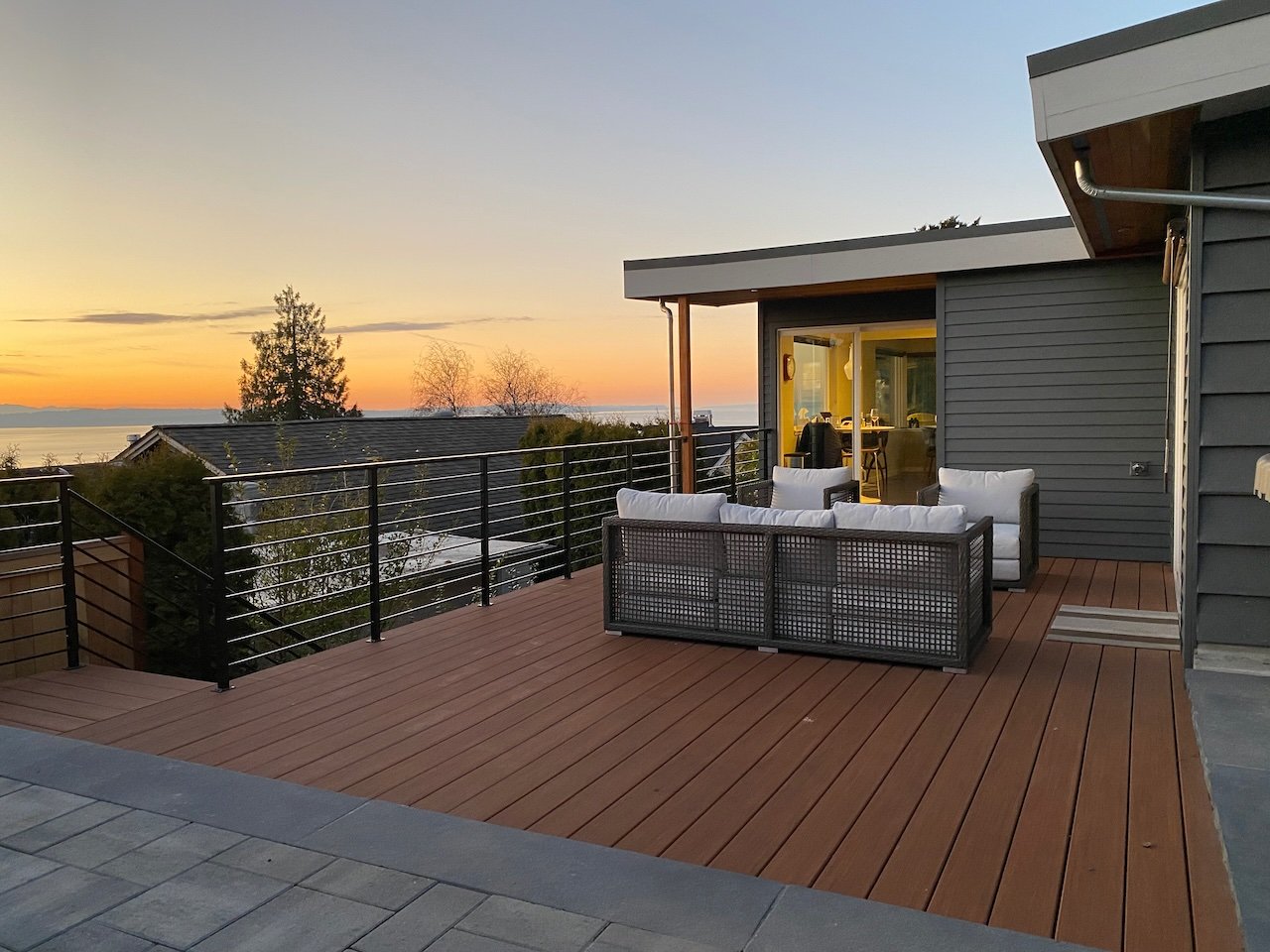 Modern rooftop deck with outdoor seating, overlooking a sunset over the water, with trees in the distance.