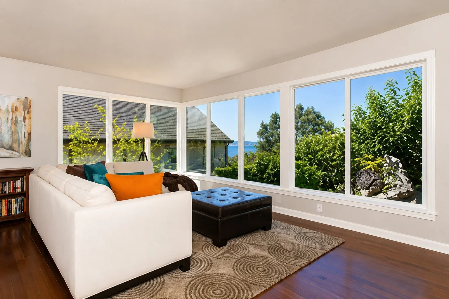 Living room with white sofa, colorful pillows, large bay windows with a view of trees and a body of water, wooden floor, area rug, and a bookshelf.