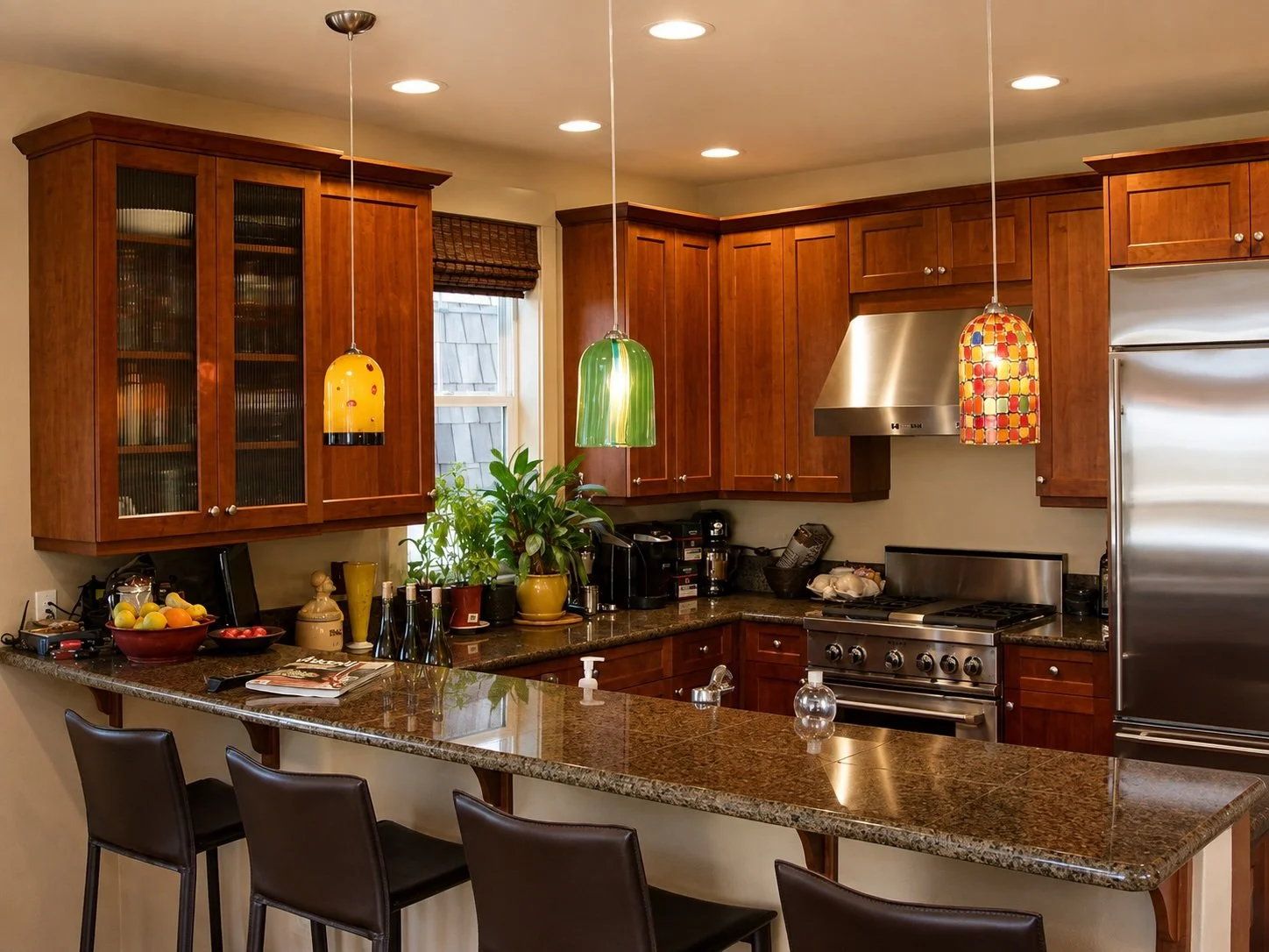 Kitchen with wooden cabinets, granite countertop, stainless steel stove and refrigerator, colorful hanging pendant lights, potted plants, fruit bowl, coffee maker, and black bar stools.