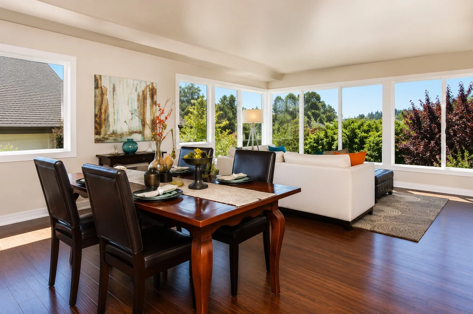Open living and dining area with large windows showing green trees outside, a dark wood dining table with black chairs, a white sofa with colorful pillows, and decorative vases and artwork.