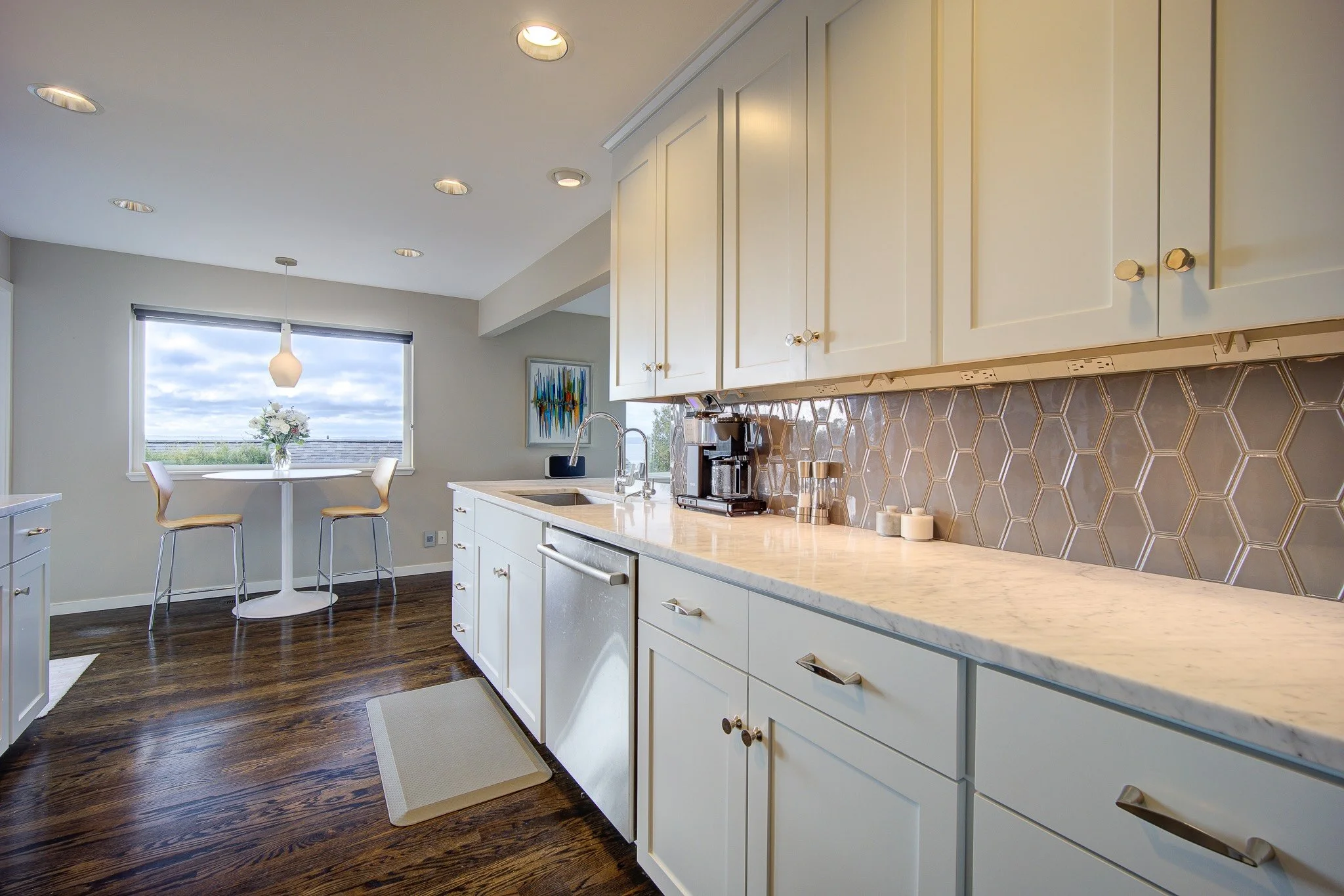 Modern kitchen with white cabinets, a marble countertop, a honeycomb tile backsplash, coffee maker, and a window overlooking a cloudy sky.