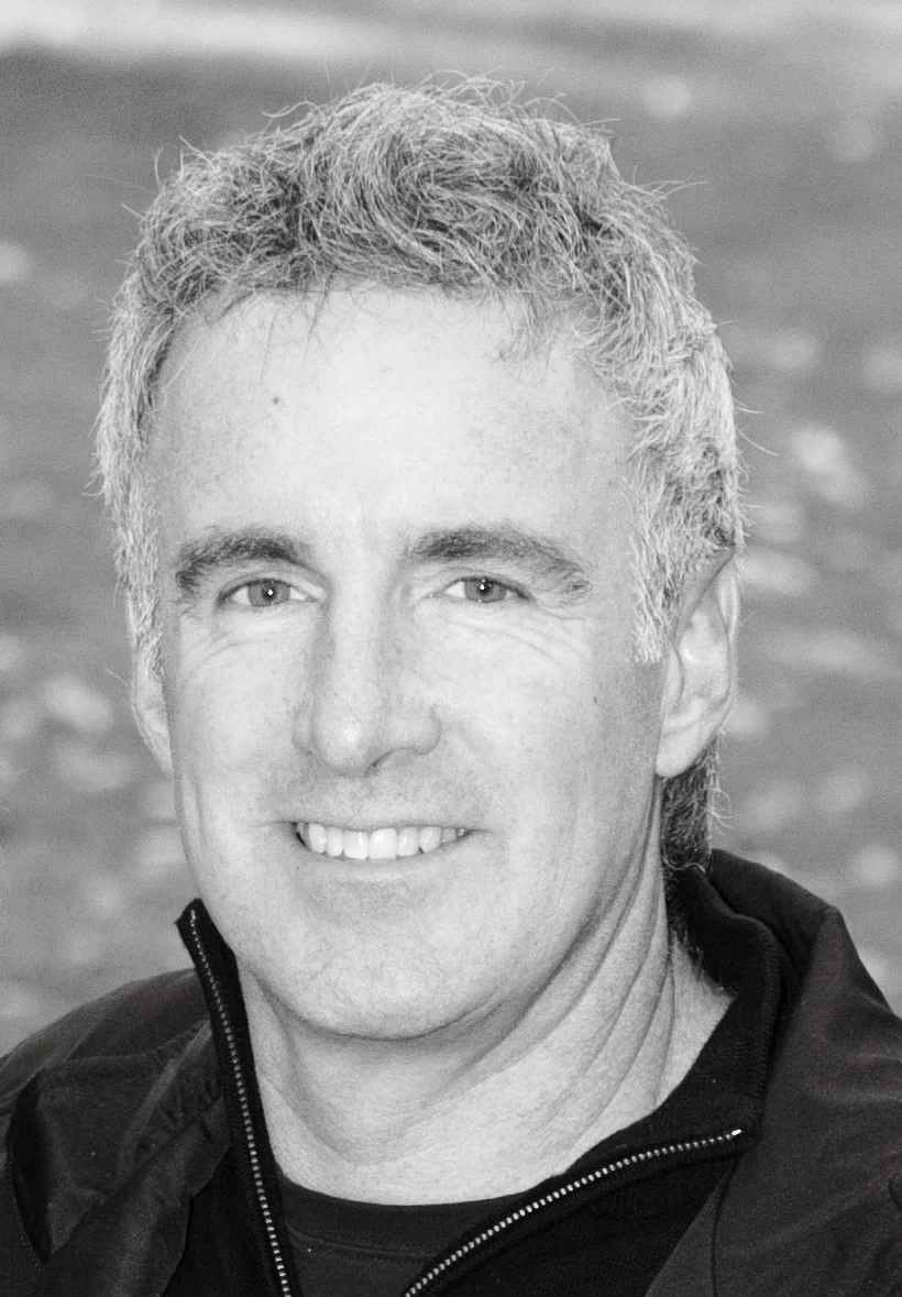 Close-up black and white photo of a smiling man with short, curly gray hair, wearing a dark jacket.