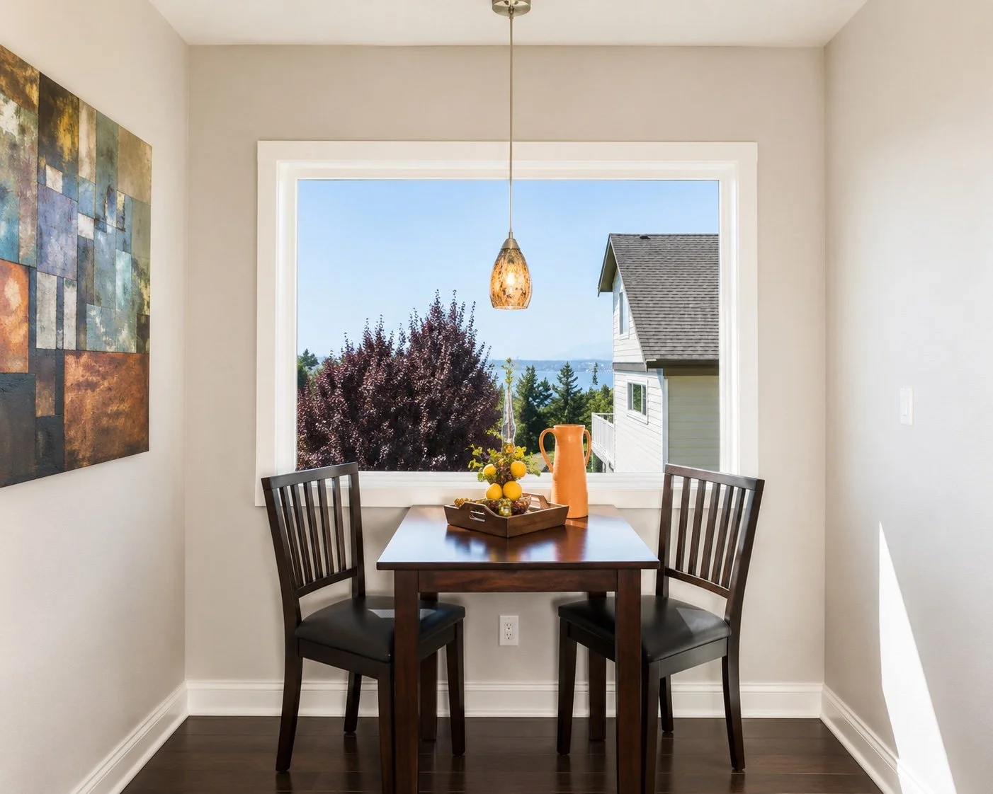 A small dining area with a wooden table and two black chairs, placed by a large window with a view of trees and neighboring houses. On the table, there is a tray with yellow lemons and a peach-colored pitcher, with a hanging pendant light above.
