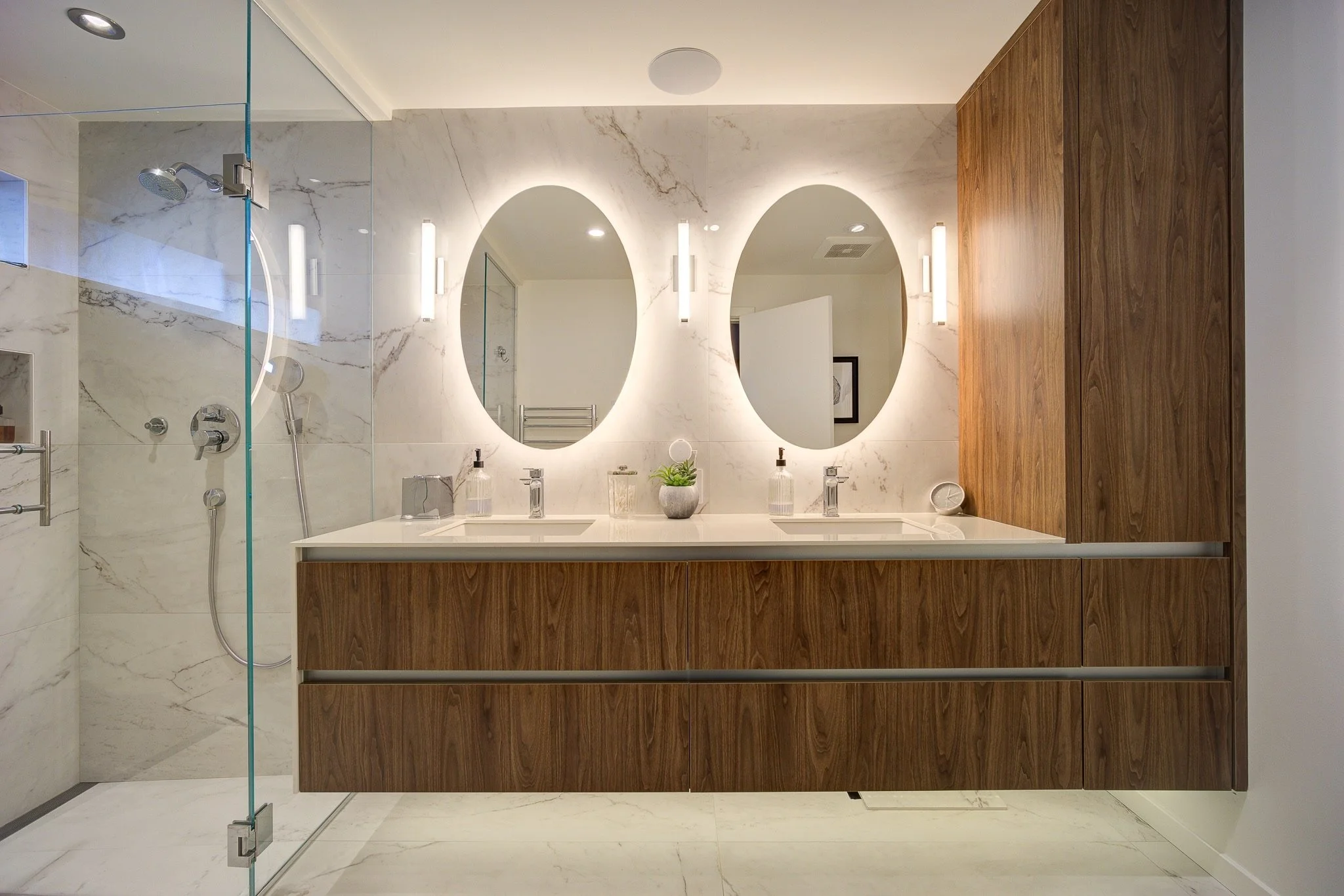 Modern bathroom with marble walls and floor, featuring two oval mirrors above a double sink vanity with wooden cabinet. There is a glass-enclosed shower on the left with silver fixtures and a handheld showerhead.