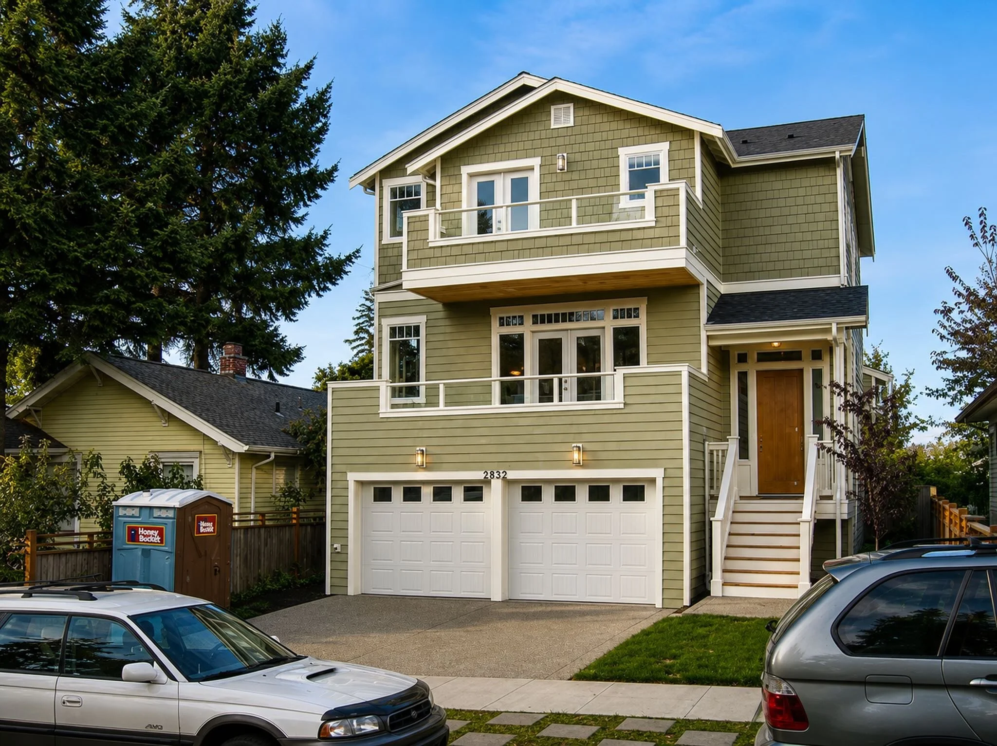A three-story green house with white trim and a garage, with cars parked in the driveway, trees around, and a blue sky overhead.