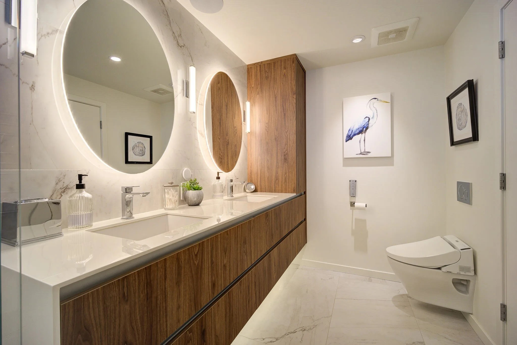 A modern bathroom with a white marble countertop, two oval backlit mirrors, wooden cabinetry, and wall art of a heron and a tree cross-section. There are soap dispensers, potted plants, a toilet, and a vent on the ceiling.