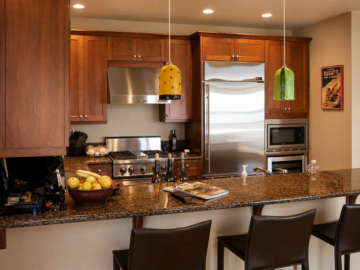 Kitchen with wooden cabinets, stainless steel appliances, a granite countertop island, and three black chairs. Items on the counter include apples, bottles, a magazine, and kitchen tools. Two colorful hanging pendant lights are above the island. Ther