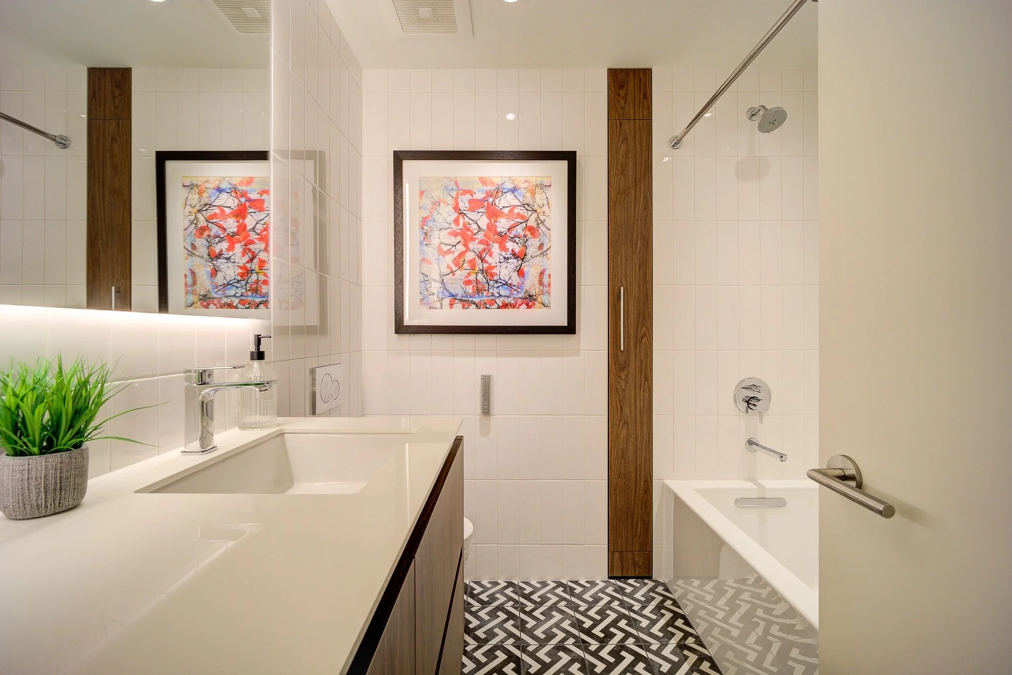 A modern bathroom with white tiled walls, two framed colorful paintings of red and purple leaves, a large mirror, a white sink with a green plant, and a shower area with a wooden panel, a shower head, and patterned floor tiles.