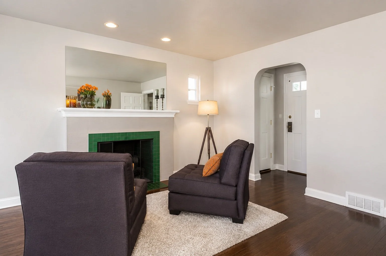 Living room with dark gray chairs, a fireplace with green tiles, a beige area rug, a tall floor lamp, and decorative vases with orange flowers on the mantel.
