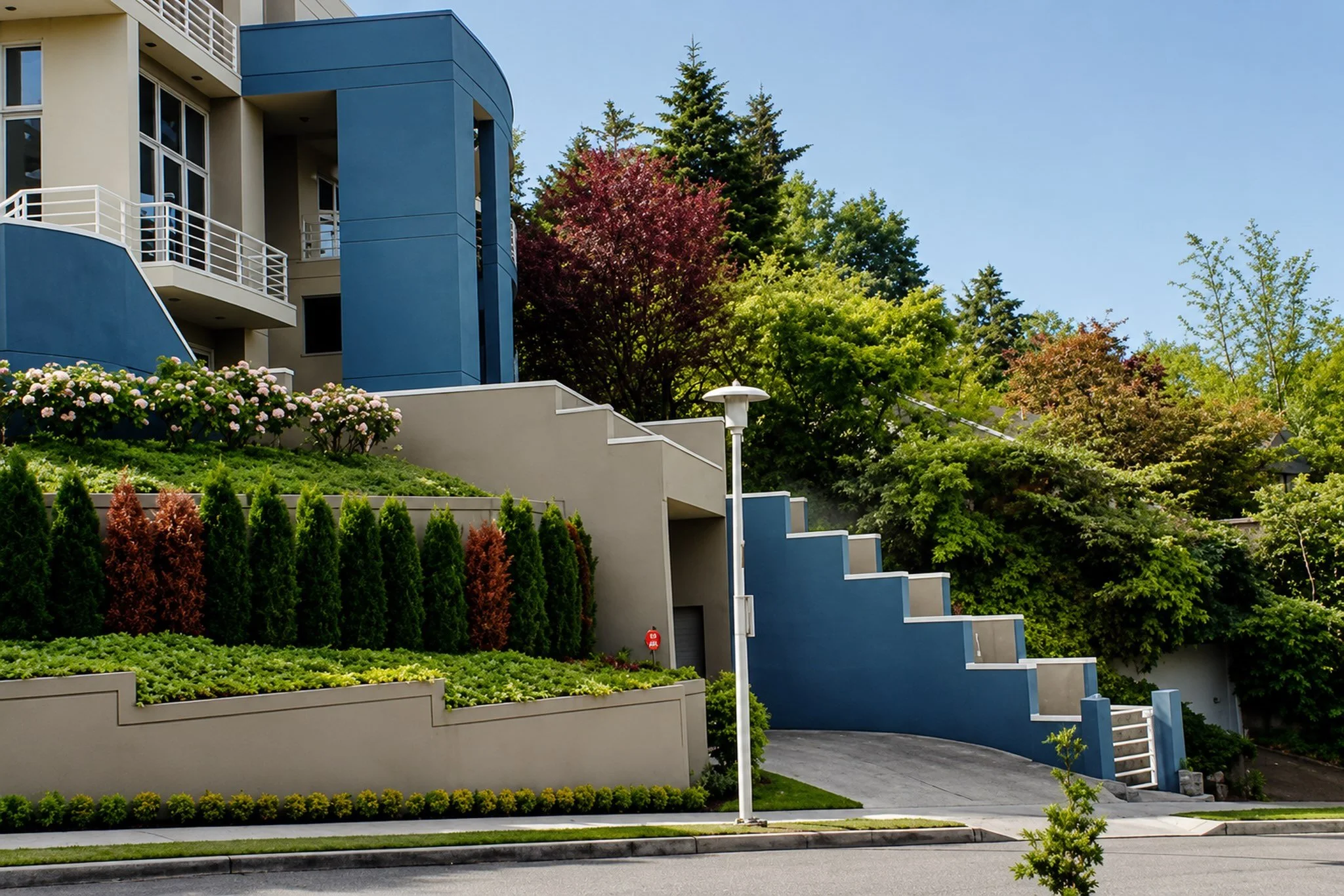 Multi-story modern residential buildings with blue and beige facades, surrounded by landscaped gardens with pink, green, and red plants, and tall trees in the background under a clear blue sky.