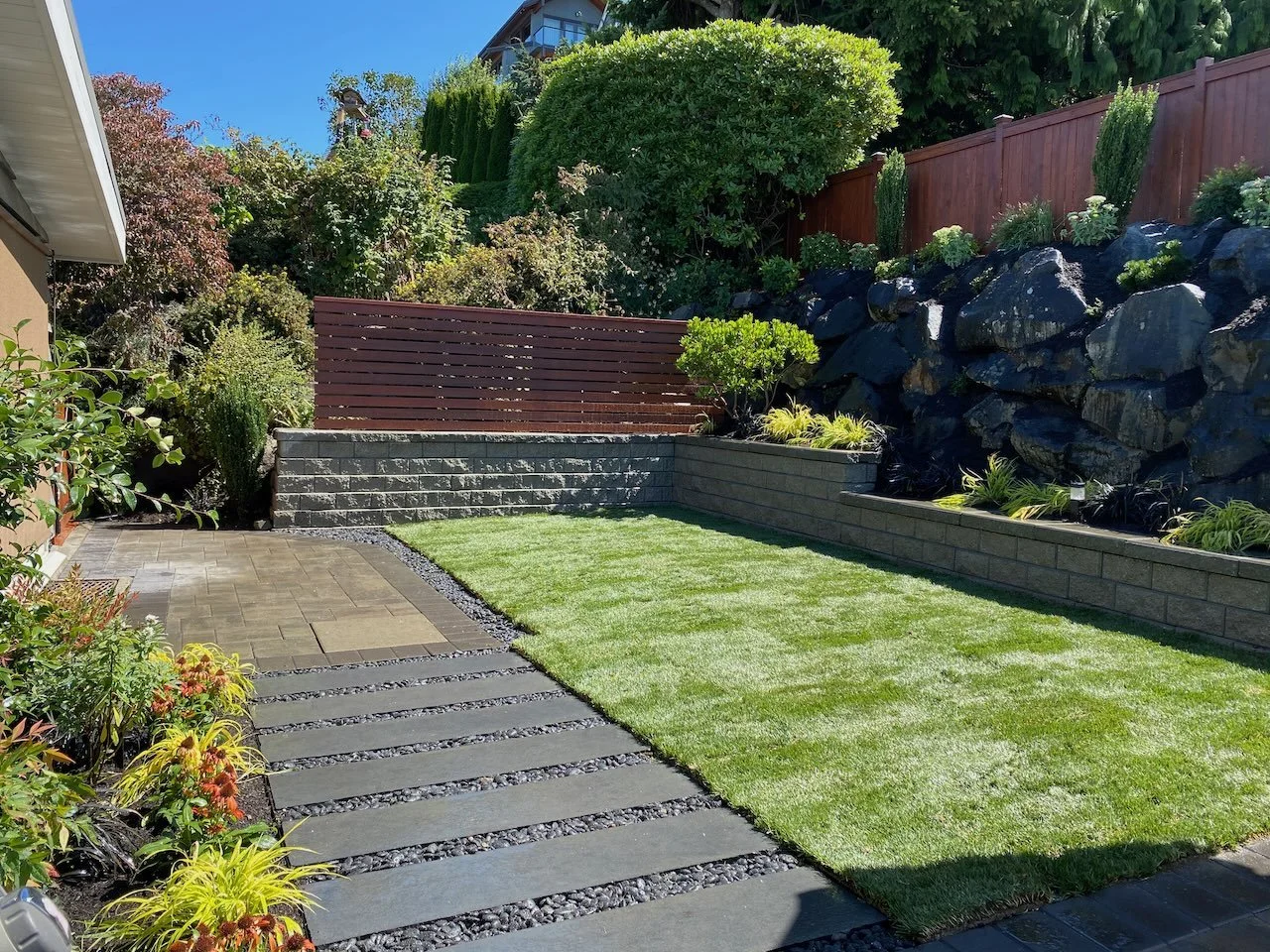 A backyard with a small patch of green grass, a stone pathway, a stone retaining wall, and garden beds with shrubs and flowers, bordered by a wooden fence and a large rock formation.