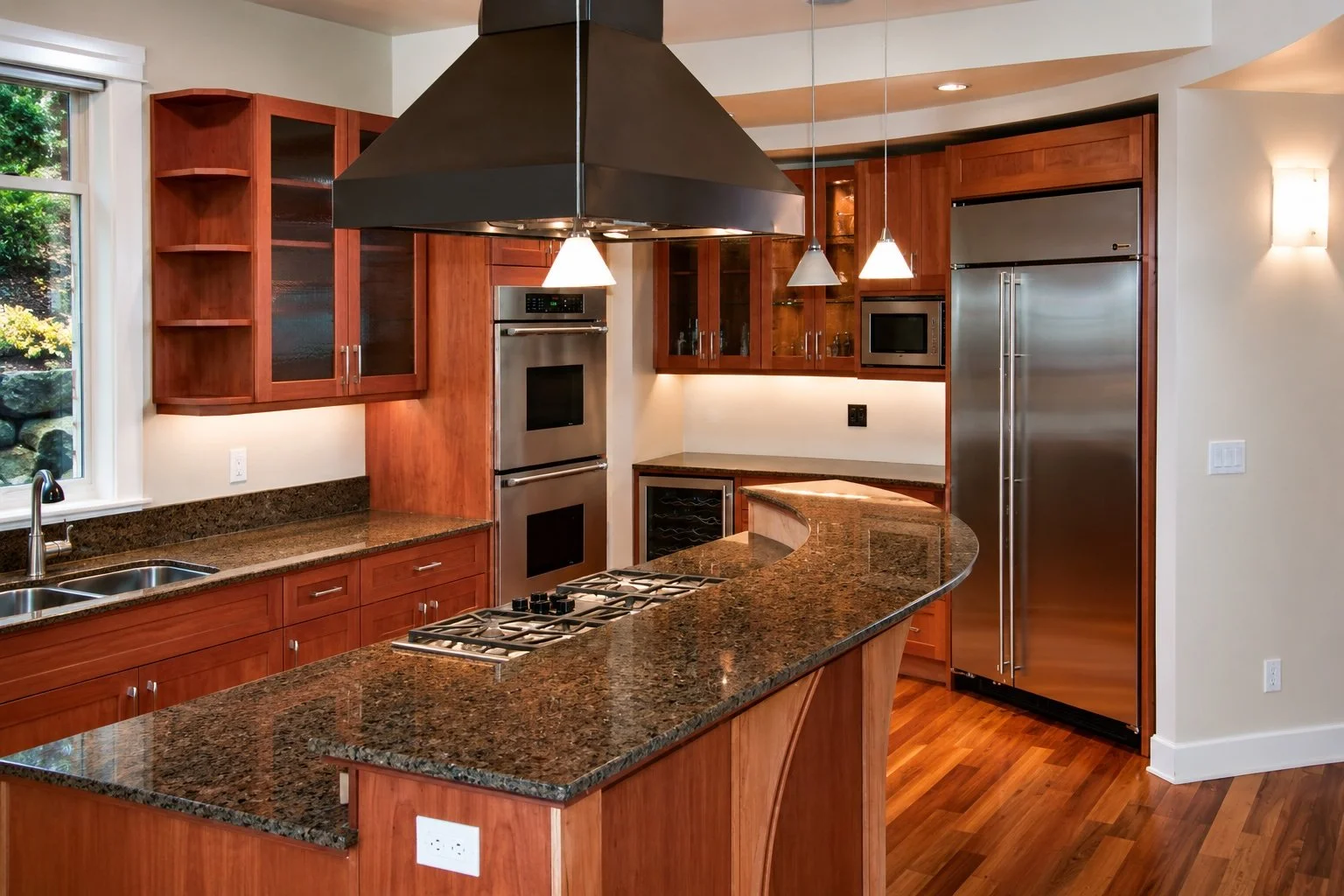 Modern kitchen with wooden cabinets, granite countertops, a large stainless steel refrigerator, a built-in microwave, double wall ovens, and a gas stove on an island. There is a window with a view of greenery outside, and pendant and wall lighting.