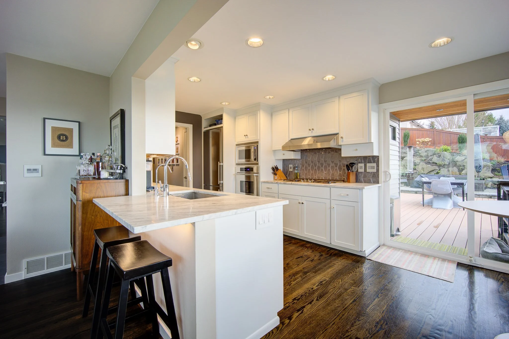 Bright kitchen with white cabinets, stainless steel appliances, a marble countertop island, dark wood floors, and sliding glass door leading to an outdoor patio with deck furniture and a landscaped yard.