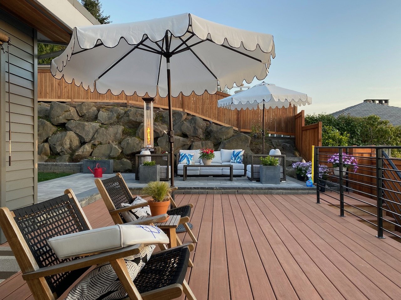 An outdoor patio with wooden decking, white umbrellas, a lounge area with cushions, potted plants, and flowers, against a rocky hillside with a wooden fence and neighboring houses in the background during daytime.