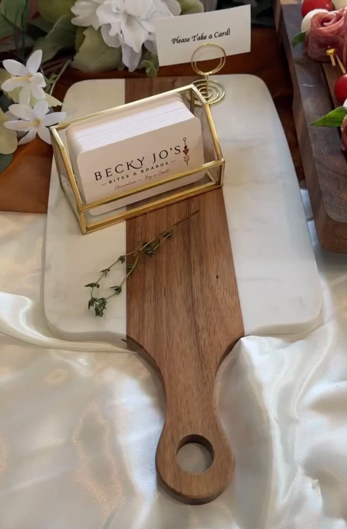 A wooden paddle with a hole at the handle's end, used for serving food, with a display of business cards for Becky Jo's Bites & Boards in a gold frame holder on a table decorated with flowers and a sign that reads "Please Take a Card."