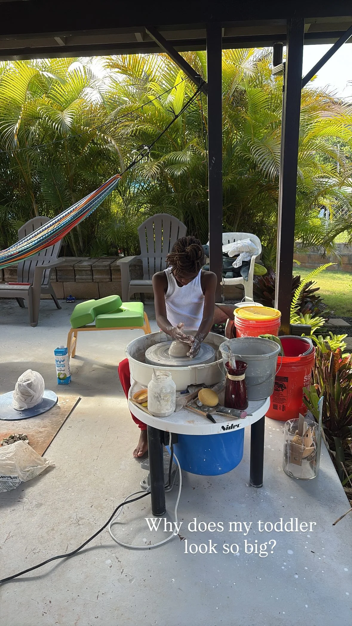 A woman with dreadlocks working on a pottery wheel outdoors with lush green plants in the background. Various pottery supplies and tools are on the table and surrounding area. The caption reads: 'Why does my toddler look so big?'