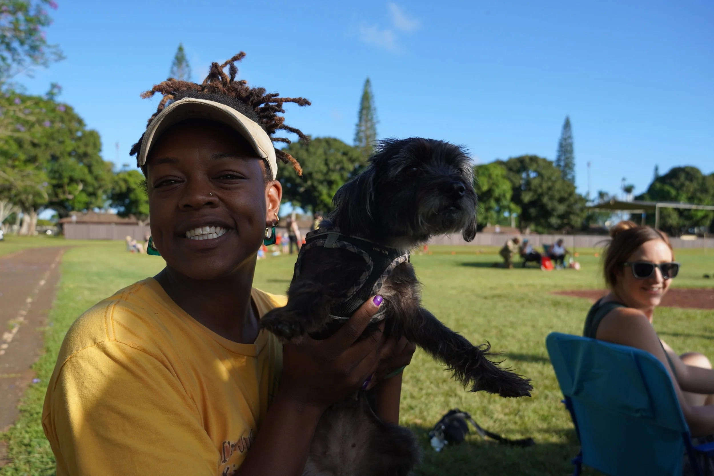 A person smiling while holding a small black dog outdoors in a park with green trees and people sitting on benches in the background.